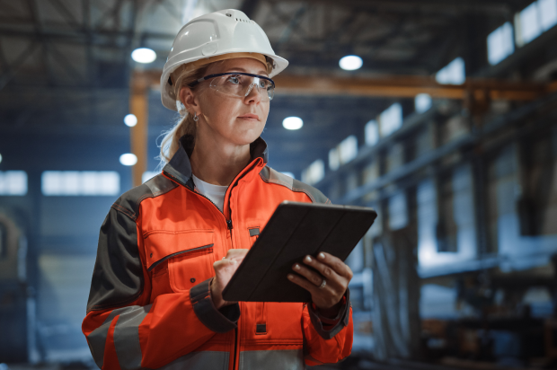 Manufacturing worker wearing a hardhat and orange safety jacket reviewing something on a tablet
