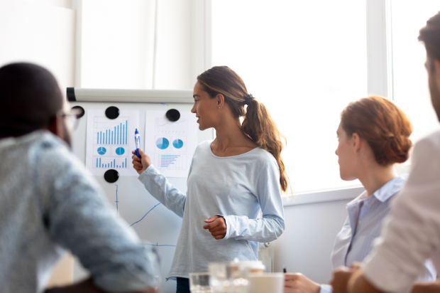 Female pointing to graphs on a whiteboard in a meeting room with coworkers