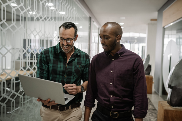 Two coworkers walking through an office setting hallway discussing something on a laptop