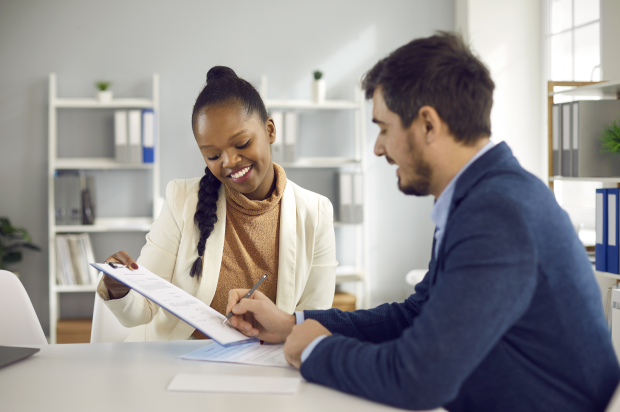 Two coworkers sitting at a desk with one coworker signing a document on a clipboard