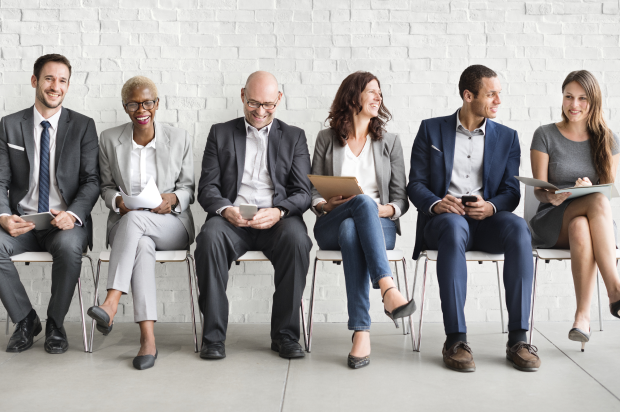 Group of smiling people sitting in a line of chairs, all dressed business smart