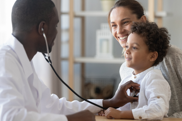 Male doctor using a stethoscope to check a young boy’s heartbeat. Young boy is sitting on his mother’s lap