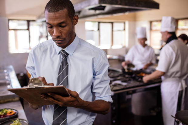 Male in a restaurant kitchen taking notes on a clipboard with chefs cooking in the background