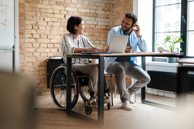 Two smiling coworkers sitting at a desk discussing something on a laptop